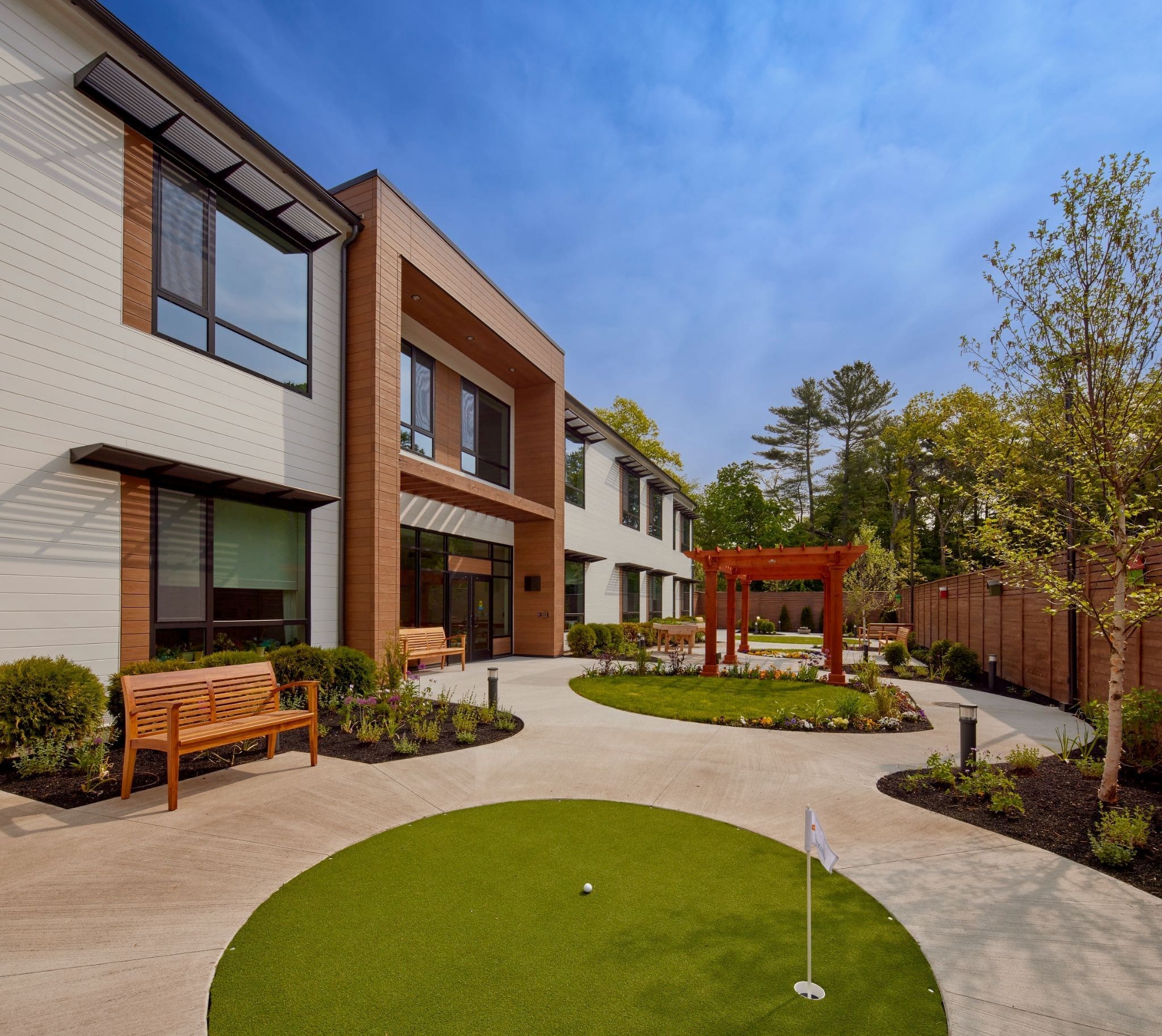 Modern building courtyard with benches, a small putting green, landscaped gardens, a wooden pergola, and trees under a blue sky.