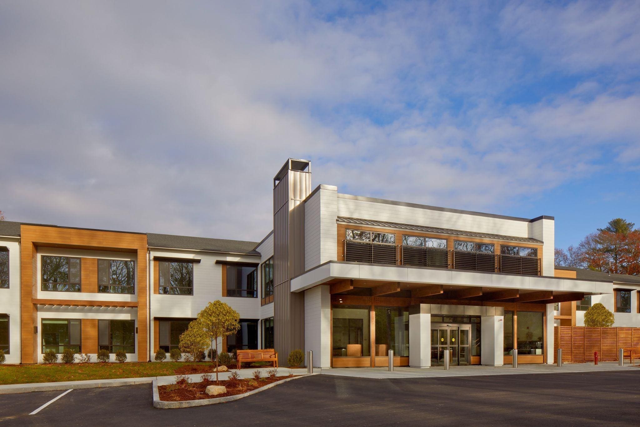 Modern two-story building with large windows, wood and white exterior accents, glass entrances, and landscaped surroundings under a partly cloudy sky.