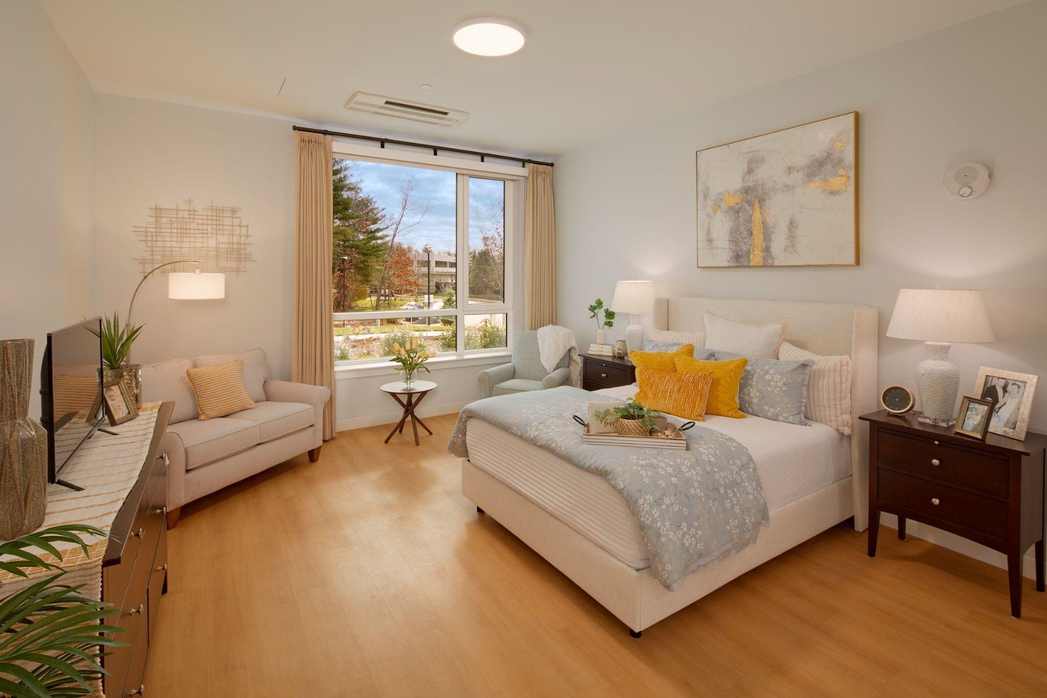 Bright, modern bedroom with a large window, bed with yellow and gray pillows, side tables with lamps, a cozy loveseat, indoor plants, and minimalist decor on light wood floors.