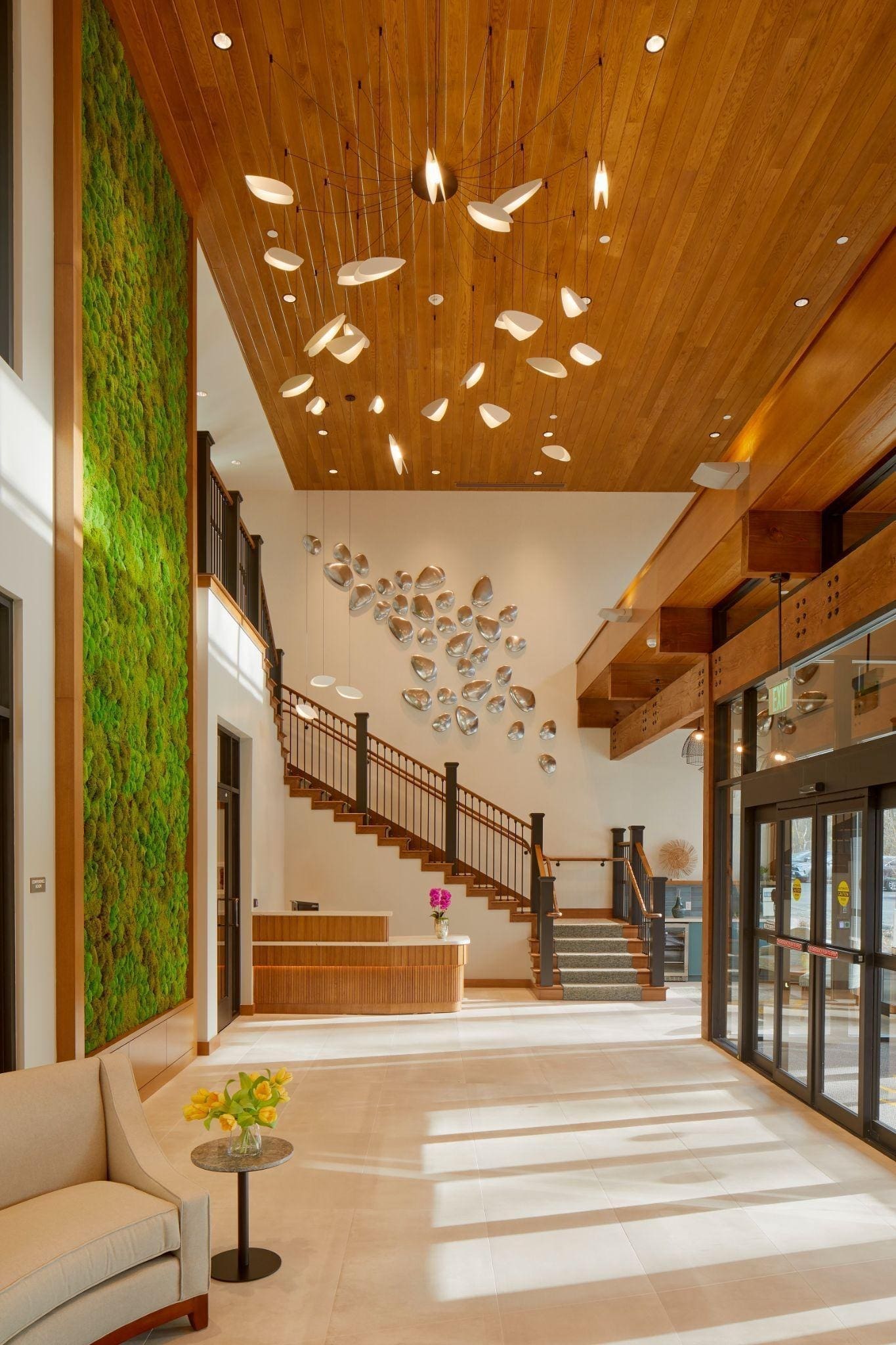 Modern lobby with a green living wall, wooden ceiling, artistic light fixture, staircase with glass and wood railing, and a glass entrance. A curved sofa and small table with yellow flowers are in the foreground.