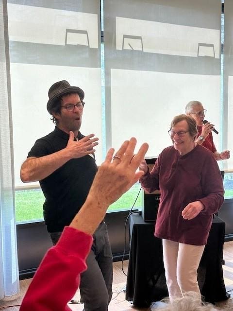 A man in a hat and a woman smile and dance together indoors as another person sings into a microphone. An arm is raised in the foreground.