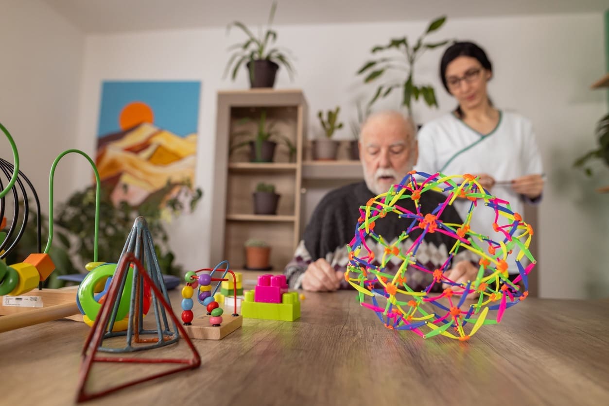 An elderly man sits at a table with colorful geometric toys, focusing on the objects, while a caregiver stands nearby in a bright room with potted plants and artwork on the wall.