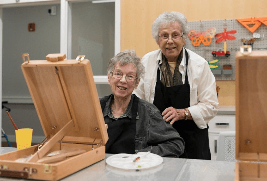 Two elderly women smile in an art room, one seated at a table with art supplies and an easel, the other standing beside her. Both wear black aprons, enjoying colorful crafts and the benefits of Memory Care with Music Therapy.