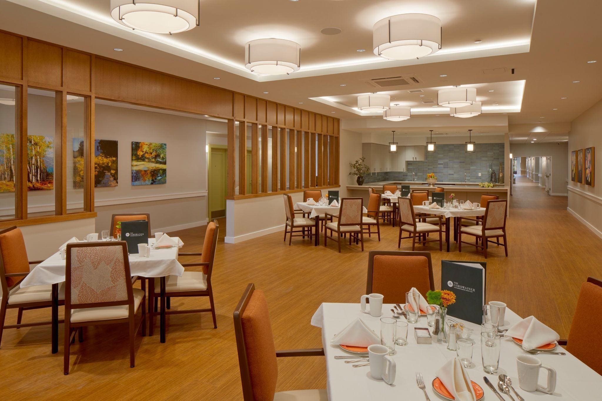 A spacious, modern dining room with round tables set for meals, cushioned chairs, warm wood flooring, contemporary ceiling lights, and colorful landscape artwork on the walls.