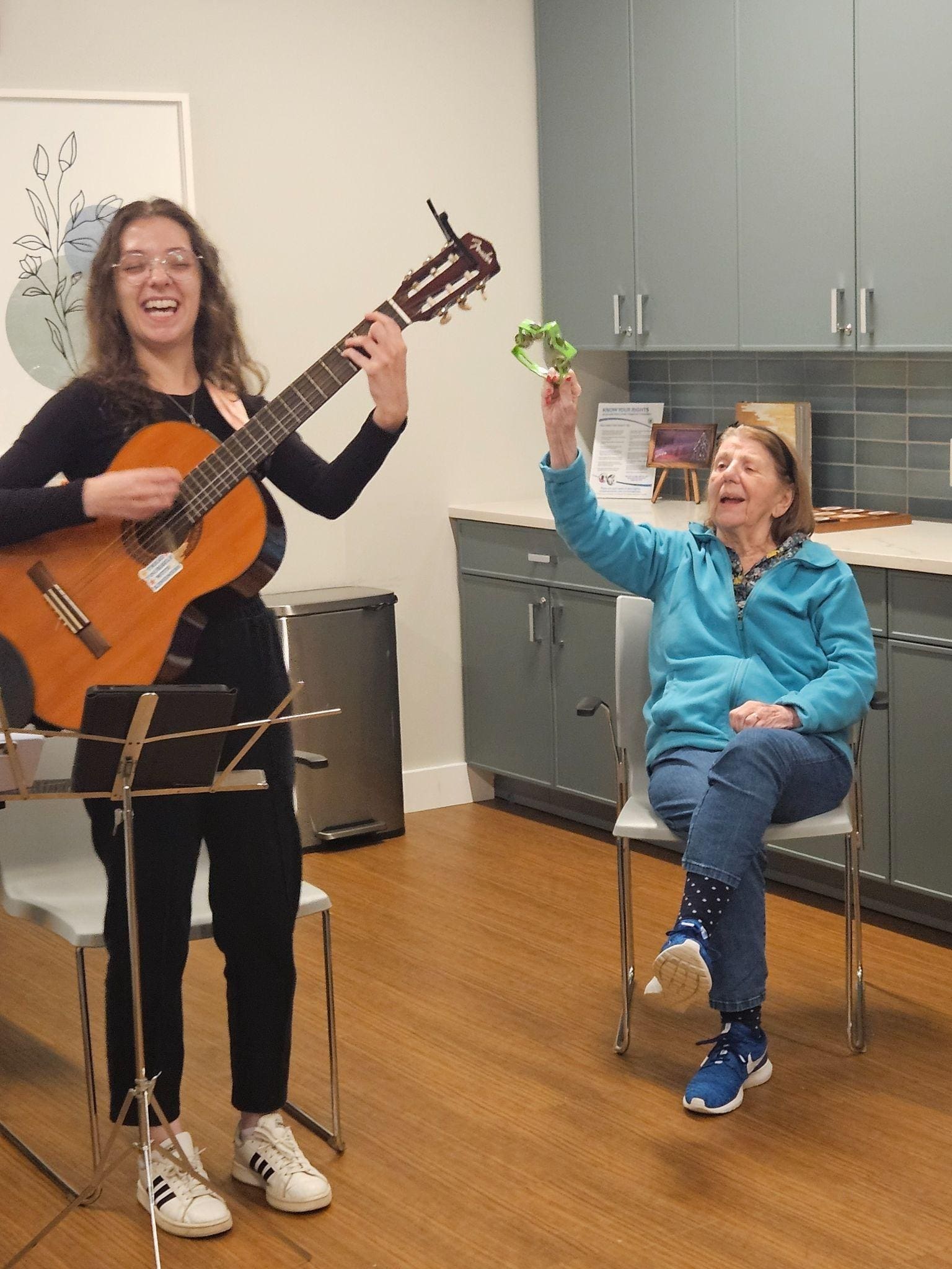 A young woman plays guitar and sings while an older woman sitting nearby smiles and shakes a tambourine in a cheerful indoor setting.