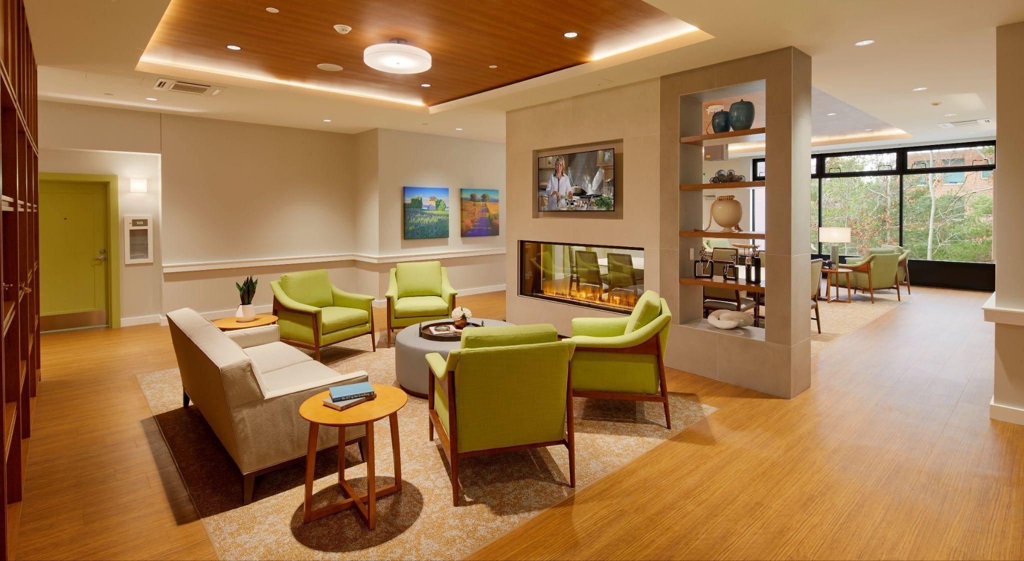 Modern lounge with green chairs, beige sofas, a double-sided fireplace, wooden floors, and shelves with decor. Large windows let in natural light; paintings and books add to the cozy ambiance.