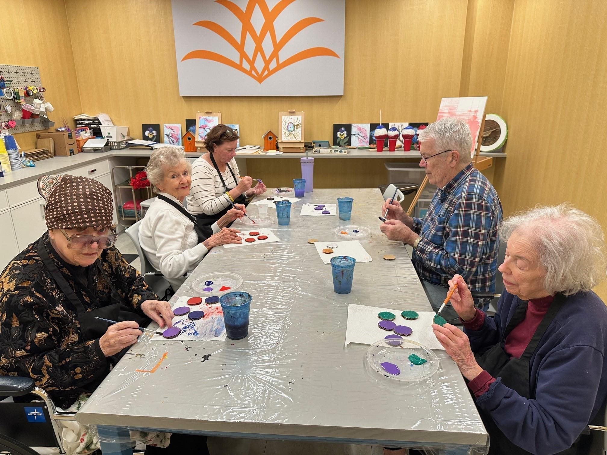 Five seniors sit at a table painting colorful circles on paper in an art room. Paints, brushes, and cups are on the table. Artworks are displayed on a counter and wall behind them.
