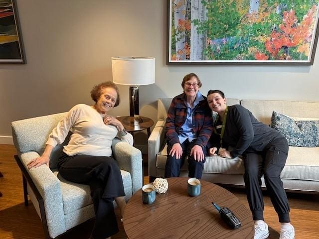 Three women sit and smile together in a living room. Two are on a couch, one is on a chair. A lamp and colorful artwork are behind them, with mugs and a radio on the coffee table in front.