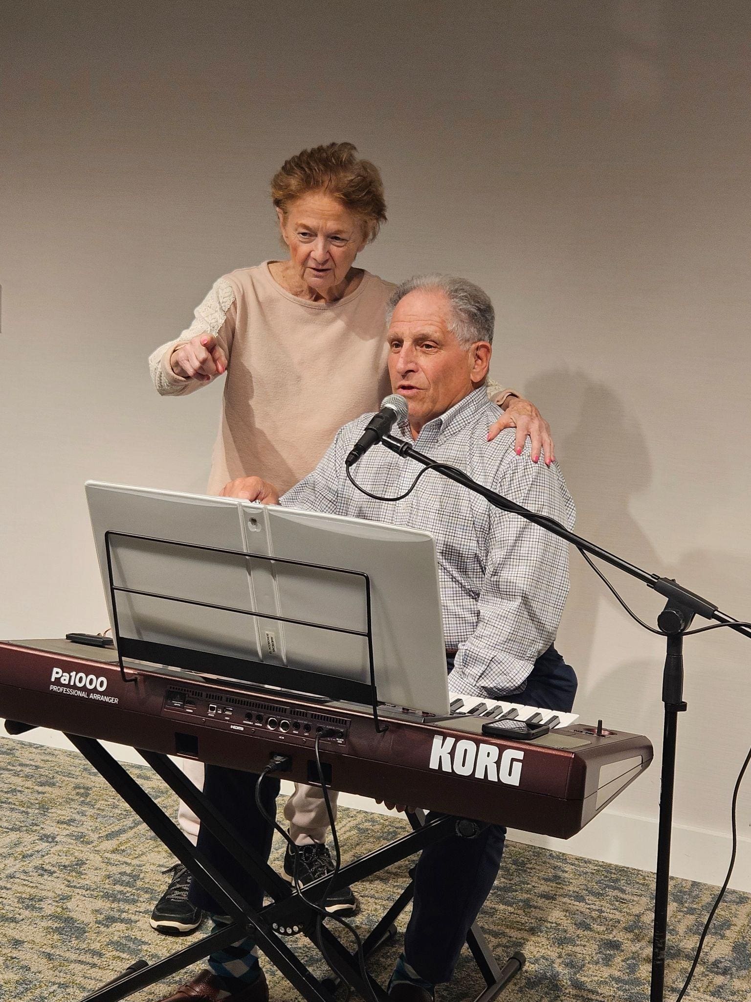 An older man plays a Korg keyboard and sings into a microphone. An older woman stands beside him with one hand on his shoulder, pointing forward. Both appear focused and engaged in the musical performance.