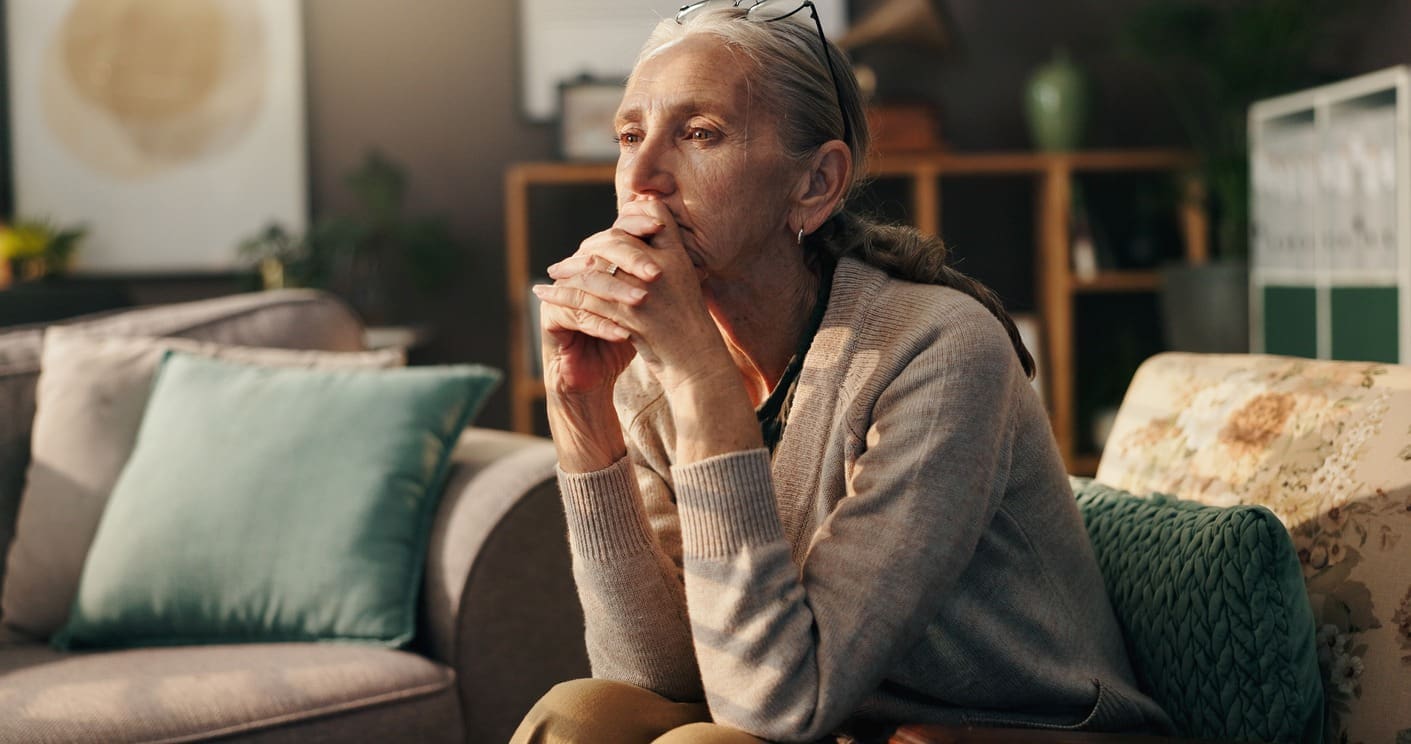 An older woman sits on a couch with her hands clasped in front of her face, looking thoughtful and concerned. She wears glasses on her head and a beige cardigan. The room is softly lit and cozy.