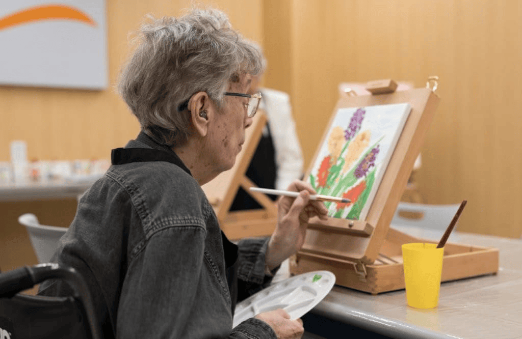 An older person with gray hair and glasses sits in a wheelchair, painting colorful flowers on a canvas at an art easel—a joyful moment in Memory Care on the South Shore MA. Art supplies and a yellow cup are on the table in front of them.
