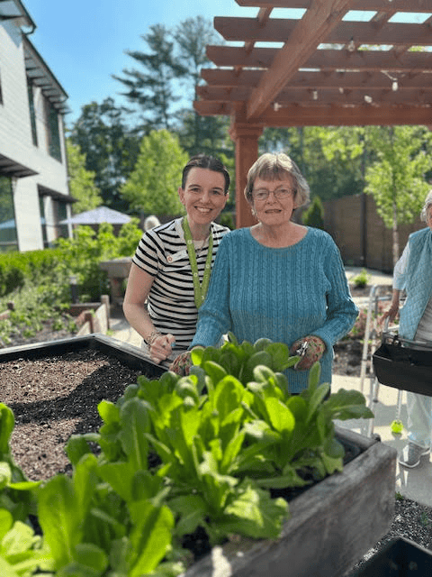 Two women, one older wearing a blue sweater and one younger in a striped shirt, smile together while gardening outdoors near a raised planter box with green plants. Sunlight and trees are visible in the background.