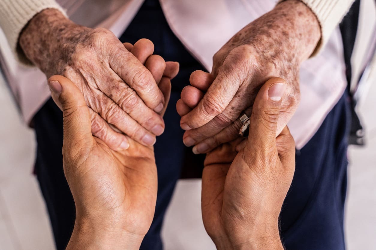 Close-up of two younger hands gently holding the hands of an elderly person, conveying care, support, and connection. The elderly person wears a light sweater and has visible age spots on their hands.