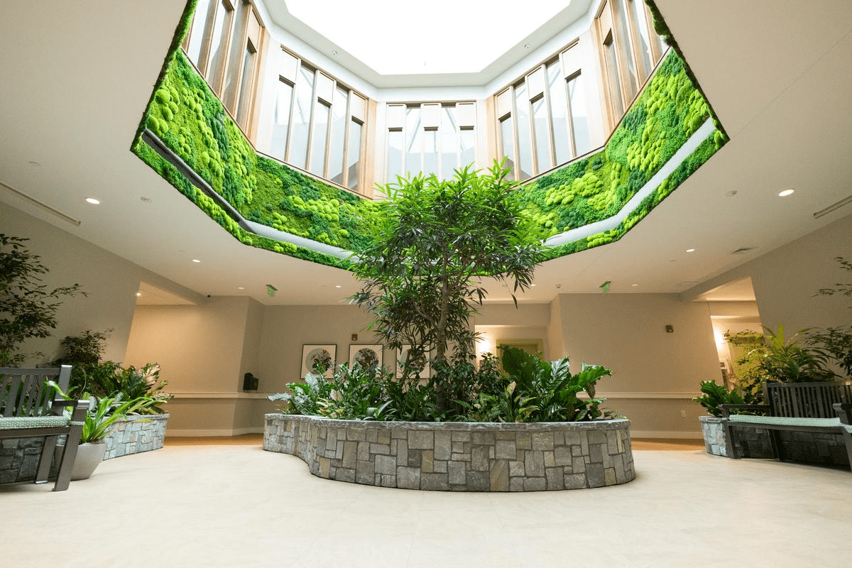 A bright indoor atrium with a skylight at The Cordwainer, featuring a central stone planter with lush green plants and a tree, surrounded by benches and accented with vertical living plant panels on the upper walls.