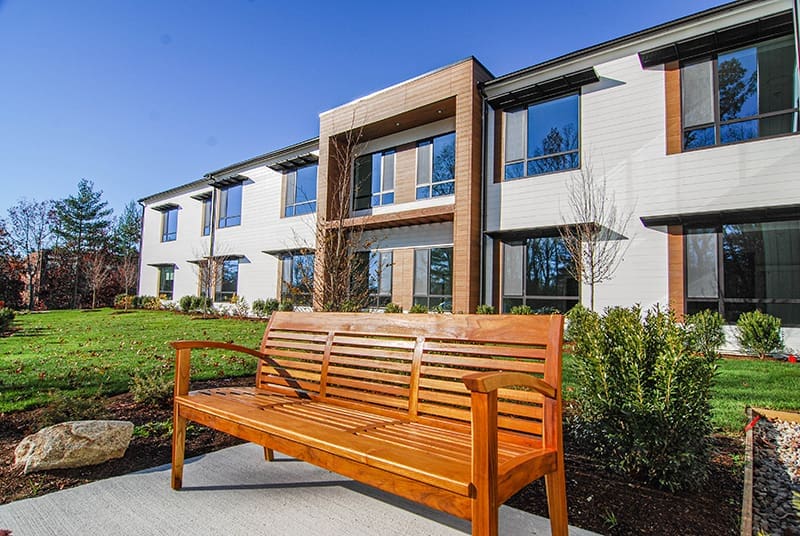 A wooden bench sits on a concrete path in front of a modern two-story building with large windows, designed as an Alzheimer's care environment, surrounded by green grass, shrubs, and small trees under a clear blue sky.