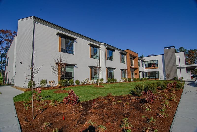 The Cordwainer's exterior in Norwell, MA, a modern two-story building with large windows and warm white and brown walls, surrounded by landscaped gardens and young trees.