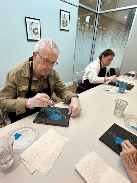 Two elderly people sit at a table in a memory care Boston community, focused on painting with blue paint on black canvases. Art supplies, water cups, and paper towels fill the table. Framed artwork hangs on the glass wall behind them.